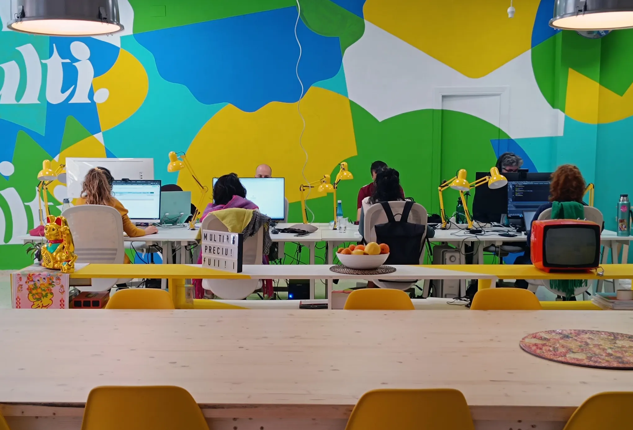 Personas trabajando en Multiprecio, en mesas blancas con flexos amarillos, frente a un gran mural verde, azul y amarillo